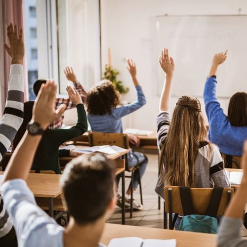 Rear view of large group of students raising their hands to answer the question on a class.