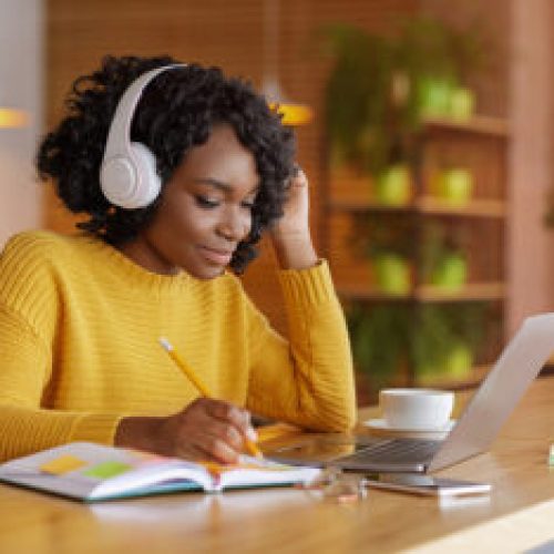 Smiling black girl with headset studying online, using laptop Smiling black girl with wireless headset studying online, using laptop at cafe, taking notes, copy space